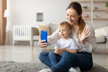 Bir anne ve küçük oğlu, doğal ışıkla dolu sıcak bir oturma odasında görüntülü bir konuşma yapıyorlar. Yumuşak bir halıda otururken gülümser ve etkileşirler..
