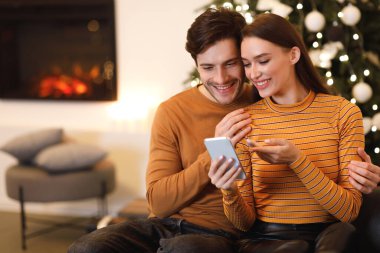 Bir çift birbirine yakın oturuyor, gülümsüyor ve akıllı telefona bakarken bir anı paylaşıyorlar. Şöminenin sıcaklığı ve tatil süslemeleri şenlikli bir atmosfer yaratıyor..