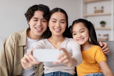 Genç bir aile rahat bir kanepede yan yana oturur. Selfie çekerken gülümserler. Parlak oda yumuşak mobilyalara sahip, sıcak ve davetkar bir atmosfer yaratıyor..