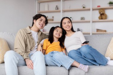 Üç aile üyesi kanepede rahatça oturur, gülümser ve film gecesinin tadını çıkarırlar. Oturma odasında hafif ışıklandırma ve neşeli bir ortam var..