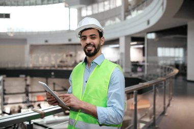 İyi aydınlatılmış bir binada güvenlik yeleği ve kask giyen inşaat işçisi inşaat görevlerini izlemek ve ekiple iletişim kurmak için tablet kullanıyor..