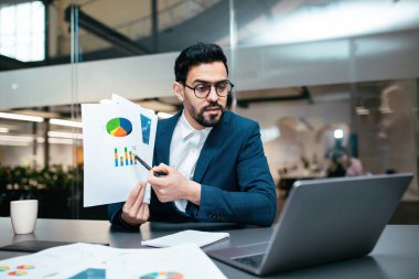 Masada oturan takım elbiseli bir adam çizelgeleri ve grafikleri sergiliyor. Önemli istatistikleri vurgulamak için bir kalem kullanıyor, bir iş görüşmesine katılıyor. Bir laptop ve kahve fincanı mevcut..