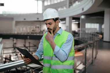 Yeşil güvenlik yeleği ve miğferi olan bir inşaat işçisi modern bir binada dururken proje planlarını inceliyor. Güneş ışığı boşluğu doldurur, detaylara odaklanır.