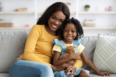 Bir anne ve kızı kanepede birlikte otururken gülümsüyorlar. Kız, sıcak oturma odalarının keyifli atmosferinin tadını çıkarırken yumuşak bir oyuncak ayı tutuyor..