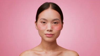 Genç bir Asyalı kadın gözlerinin altına şımartıcı cilt bakım bantları yapıştırıyor. Canlı pembe bir arka plana karşı yarı çıplak, rahatlama ve güzelliği somutlaştırıyor..