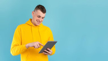 Parlak sarı kapüşonlu bir adam tabletle iletişim kurarken gülümsüyor. Arka plan koyu mavi, cihaza bağlandığı zaman neşeli yüz ifadesini vurguluyor..