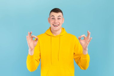 Parlak sarı kapüşonlu genç bir adam mavi bir arka plana karşı duruyor, iki eliyle iyi bir jest yaparken geniş bir gülümseme sergiliyor. Yüzü neşeli ve neşeli..