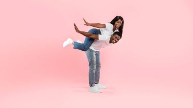 Neşeli bir Afro-Amerikan erkeği kadınını sırtında taşır, hem gülümser hem de eğlenir. Pembe bir stüdyonun önünde duruyorlar, birlikte kaygısız bir anın tadını çıkarıyorlar..