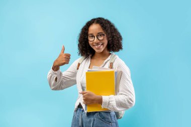 Neşeli siyah kız öğrenci notlar ve sırt çantasıyla poz veriyor. Kameraya gülümserken başparmağını kaldırıyor, modern akademik eğitimin yararlarını mavi bir zemine karşı destekliyor..