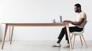 Genç siyahi bir adam evindeki bir masada oturuyor, çalışmak ya da çalışmak için dizüstü bilgisayar kullanıyor. Yakınlarda minimalist bir ortamda bir kahve fincanı ile odaklanmış durumda..