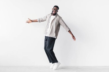 Sıradan giyinmiş neşeli genç bir siyah adam parmak uçlarında duruyor, mutluluğunu ifade ediyor ve başarısını kutluyor. Beyaz bir stüdyo duvarına karşı poz veriyor, neşeli bir atmosfer yaratıyor..