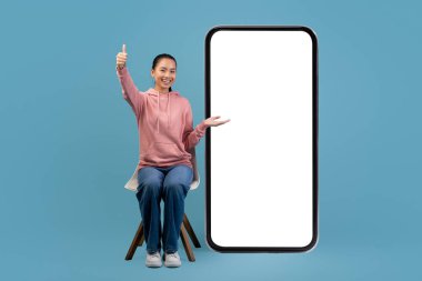 Neşeli genç bir Çinli kadın bir sandalyede oturuyor, gülümsüyor ve yanındaki boş cep telefonunu işaret ediyor. Eğlenmek için eğlenceli mobil bir uygulama öneriyor..