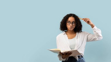 Düşünceli genç siyah bir kız öğrenci elinde kalem tutarken derin düşüncelere dalmış bir ders kitabı okuyor. Açık mavi arka planlı bir stüdyoda, sakin bir atmosfer yaratıyor..