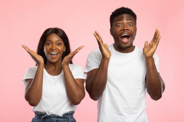 Genç siyah çift harika haberler aldıktan sonra saf heyecan ve neşe ifade ediyor. Açık ağızlarla poz veriyorlar ve ellerini kaldırıyorlar. Pembe bir arka plana karşı şaşkınlıklarını gösteriyorlar..