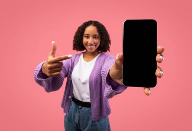 Neşeli siyah kadın cep telefonunu boş bir ekranla gösteriyor, canlı pembe stüdyo ortamında yeni bir uygulama veya web sitesini tanıtıyor. Seyircileri reklam fırsatlarını düşünmeye davet ediyor..