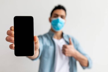 Sıradan giyinmiş genç bir adam içeride duruyor, mavi bir maske takıyor. Önünde bir akıllı telefon tutuyor ve olumlu bir deneyim ya da onayı işaret ediyor..