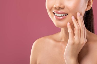 Tanımlanamayan genç bayan, mor bir arka planda kusursuz beyaz bir gülümseme sergilerken yüzüne dokunuyor. Bu kırpılmış çekim diş bakımı ve ağız hijyeninin önemini vurguluyor..