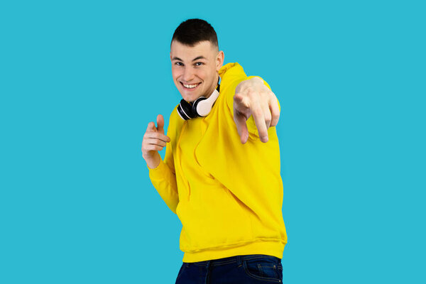 A young man smiles and points while wearing a yellow hoodie and headphones. He stands in front of a vibrant blue background, showcasing a lively and cheerful mood.