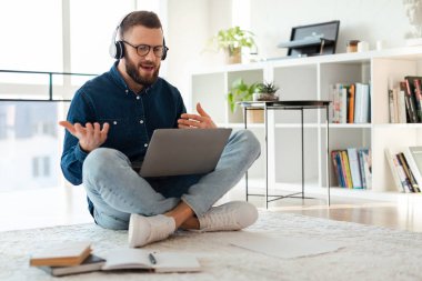 Bir adam hafif bir halıda bağdaş kurup otururken görüntülü bir görüşme yapıyor. Kulaklık takıyor ve odaklanmış görünüyor. Etrafı kitap ve bitkilerle dolu..