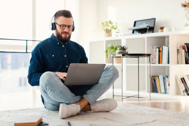 Modern bir ev ofisinde yerde oturan, kulaklık takan ve dizüstü bilgisayarda yazan bir adam. Doğal ışık içeri akar ve rahat bir çalışma alanı ortamı yaratır..