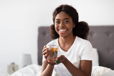 Kıvırcık saçlı neşeli bir Afro-Amerikalı kadın rahat bir şekilde yatakta oturur ve bardaktan bitki çayı yudumlar. Parlak bir tebessümle, rahat yatak odasında dinlenmenin tadını çıkarıyor..