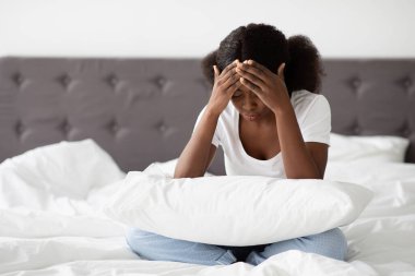 Bin yıllık siyah bir kadın pijamalarıyla yatağında oturup yastığını tutuyor ve kafasına dokunuyor. Sıkıntılı görünüyor, muhtemelen regl döneminde baş ağrısı ya da rahatsızlığı var..