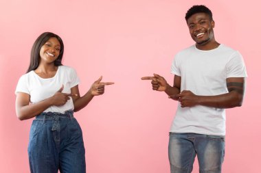 Neşeli siyah bir çift pembe bir stüdyo duvarına yaslanır, birbirlerini işaret ederken geniş bir gülümsemeyle. Günlük kıyafetleri ve enerjileri reklam afişi için eğlenceli bir ortam yaratıyor..