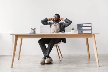 Neşeli genç siyah bir adam ahşap bir masada rahat bir şekilde oturuyor, laptopunu kullanıyor ve işe ara veriyor. Elleri başının arkasında, anın tadını çıkarıyor..