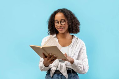 Gözlüklü siyah kız öğrenci ders kitabı okurken gülümsüyor. O parlak mavi bir stüdyo ortamında yaklaşan bir sınav için çalışıyor. Odaklanmış ifadesi öğrenmeye olan bağlılığını gösteriyor..