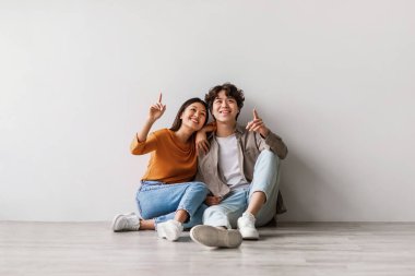 Millennial Asyalı çift beyaz bir stüdyo zemininde oturuyor, gülümsüyor ve yukarıyı işaret ediyor. Reklam tasarımı ve promosyonlar için fırsatlar yaratıyorlar, canlı ve çekici bir atmosfer yaratıyorlar..