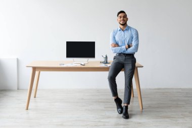 Gülümseyen bir Arap ev ofisindeki bir masada oturuyor. Kollarını kavuşturmuş ve boş siyah ekranlı bir dizüstü bilgisayar gösteriyor. Ayar parlak ve modern, uzak çalışma için ideal..