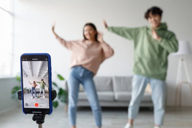 Milenyum Asyalı bir çift evde akıllı telefonlarıyla video kaydederken dans ediyor ve eğleniyor. Birlikte eğleniyorlar, rahat bir ortamda neşeli enerjilerini sergiliyorlar..