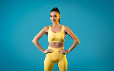 A fit woman stands with hands on hips, wearing bright yellow sportswear. She looks directly at the camera, embodying strength and confidence in a studio with a blue backdrop.