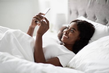 Neşeli siyah bir kadın, beyaz pijamalarıyla yatakta dinleniyor, ortağının mesajını okurken gülümsüyor. Randevu için planlar yapıyor, mutluluk ve heyecan yayıyor..