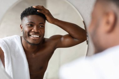 Mutlu bir Afrikalı Amerikalı duş aldıktan sonra bir banyoda üstsüz duruyor. Aynadaki yansımasına gülümserken kısa kıvırcık saçlarına dokunuyor ve sabah güzellik rutinini kucaklıyor..