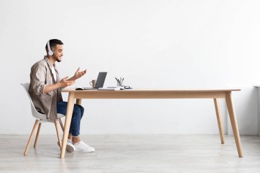 Genç bir Arap, parlak bir ofis alanındaki parlak ahşap bir masada otururken bir video görüşmesine odaklanır. Kulaklık takıyor ve uzaktan iletişim deneyiminin tadını çıkarıyor..