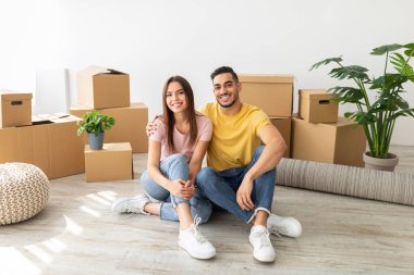 Genç bir çift yeni oturma odalarının zemininde oturuyor, kahverengi taşınan kutular ve kapalı alandaki bitkiler arasında poz verirken gülümsüyorlar. Yeni başlangıçları için heyecanlılar..