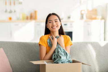 Woman sits on her couch, excitedly unboxing a delivery. She hugs a shirt, showing her satisfaction with the online purchase while smiling brightly at the camera.