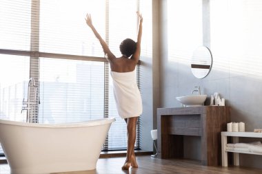 Beyaz havluya sarılı genç siyah bir bayan şık bir banyoda dans ediyor. Panoramik bir pencerenin yanında duruyor, sabah güneşinin sıcaklığını hissederken zamanının tadını çıkarıyor..