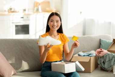 Neşeli bir Asyalı kadın kanepesinde otururken içinde beyaz spor ayakkabılar olan bir karton kutuyu açıyor. Bir kredi kartı tutuyor ve kameraya gülümsüyor, evden alışveriş deneyiminin tadını çıkarıyor..