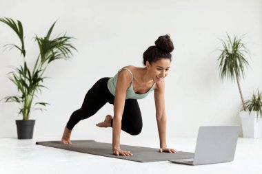 Genç bir kadın evdeki yoga minderinde spor yapıyor, bitkilerle dolu parlak bir odada dizüstü bilgisayarında video dersleri izliyor. Odaklanmış ve aktif, egzersizleri üzerinde çalışıyor..