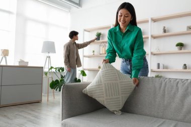 Genç Asyalı kadın rahat kanepelerine bir yastık yerleştirirken erkek arkadaşı da dekorasyonu rafa yerleştirip dairelerinin sıcak atmosferini arttırıyor. Dekorasyon sürecinden birlikte zevk alıyorlar..