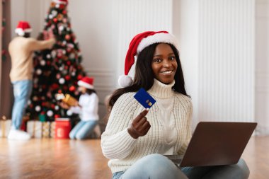 Kırmızı Noel Baba şapkalı mutlu bir milenyum hanımı internetten bilgisayarını kullanıyor ve elinde kredi kartı tutuyor. Arka planda, ailesi bir Noel ağacı süslüyordu..