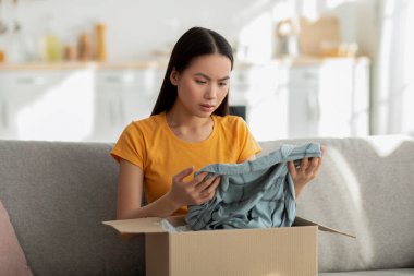 Genç bir Asyalı bayan müşteri evdeki rahat bir kanepede oturur ve içinde giysiler olan bir teslimat kutusunu açar. Ürünleri incelerken, karşılanmamış beklentilerini açığa çıkaran yüz ifadesi hayal kırıklığını gösteriyor..