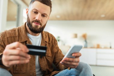 Bir adam rahat bir oturma odasında oturmuş elinde kredi kartıyla akıllı telefonuna bakıyor. Doğal ışık boşluğu doldururken çevrimiçi bir alışveriş yapmaya odaklanmış görünüyor..
