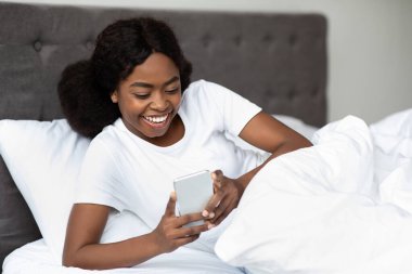 Neşeli bir milenyum siyahi bayan, akıllı telefonunu kullanarak yatakta uzanıyor. E-postaları kontrol ederken mutlu görünüyor ve sabah uyandıktan sonra arkadaşlarıyla bağlantı kuruyor..