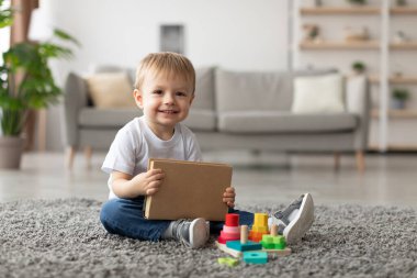 Neşeli genç bir çocuk parlak bir oturma odasında yumuşak bir halıya oturur. Kahverengi bir kitap tutuyor ve etrafında dağılmış renkli yapı taşlarıyla oynuyor, içeride geçirdiği zamanın tadını çıkarıyor..