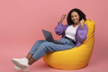 Heyecanlı genç siyah kadın parlak bir koltukta dizüstü bilgisayar kullanarak oturuyor. Kollarını zaferle kaldırıyor, canlı pembe bir arka planla online piyangodaki başarısının tadını çıkarıyor..