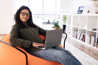 Genç bir kadın turuncu bir sandalyeye rahatça oturur ve dizüstü bilgisayarına odaklanır. Doğal ışık ve dekorla dolu şık bir oturma odasında rahat bir atmosferin tadını çıkarıyor..