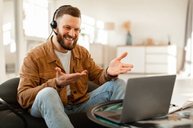 Kulaklık takan bir adam görüntülü konuşma yaparken gülümsüyor ve jestler yapıyor. Doğal ışık ve modern dekorasyonla şık bir oturma odasında rahatça oturuyor..